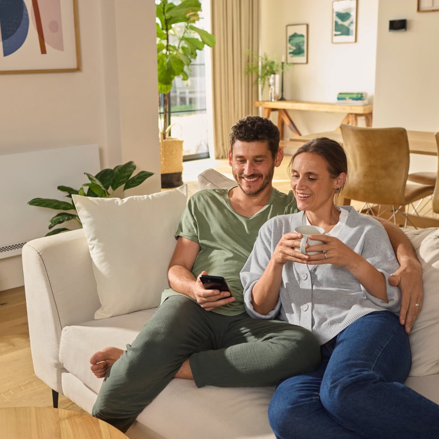 Bir çift kanepede oturmuş akıllı telefona bakıyor. Arka planda Vaillant aroVAIR plus fan coil konsol modeli görülüyor