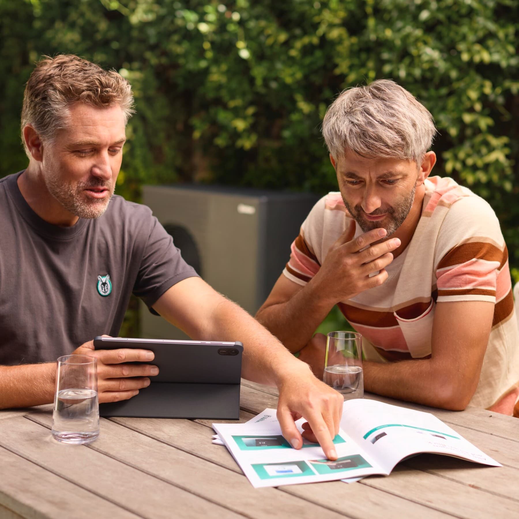 vaillant ısı pompası önünde Bir montajcı ve bir müşteri bahçede oturmuş, bir tablete bakıyorlar. Arka planda bir Vaillant ısı pompası görünüyor